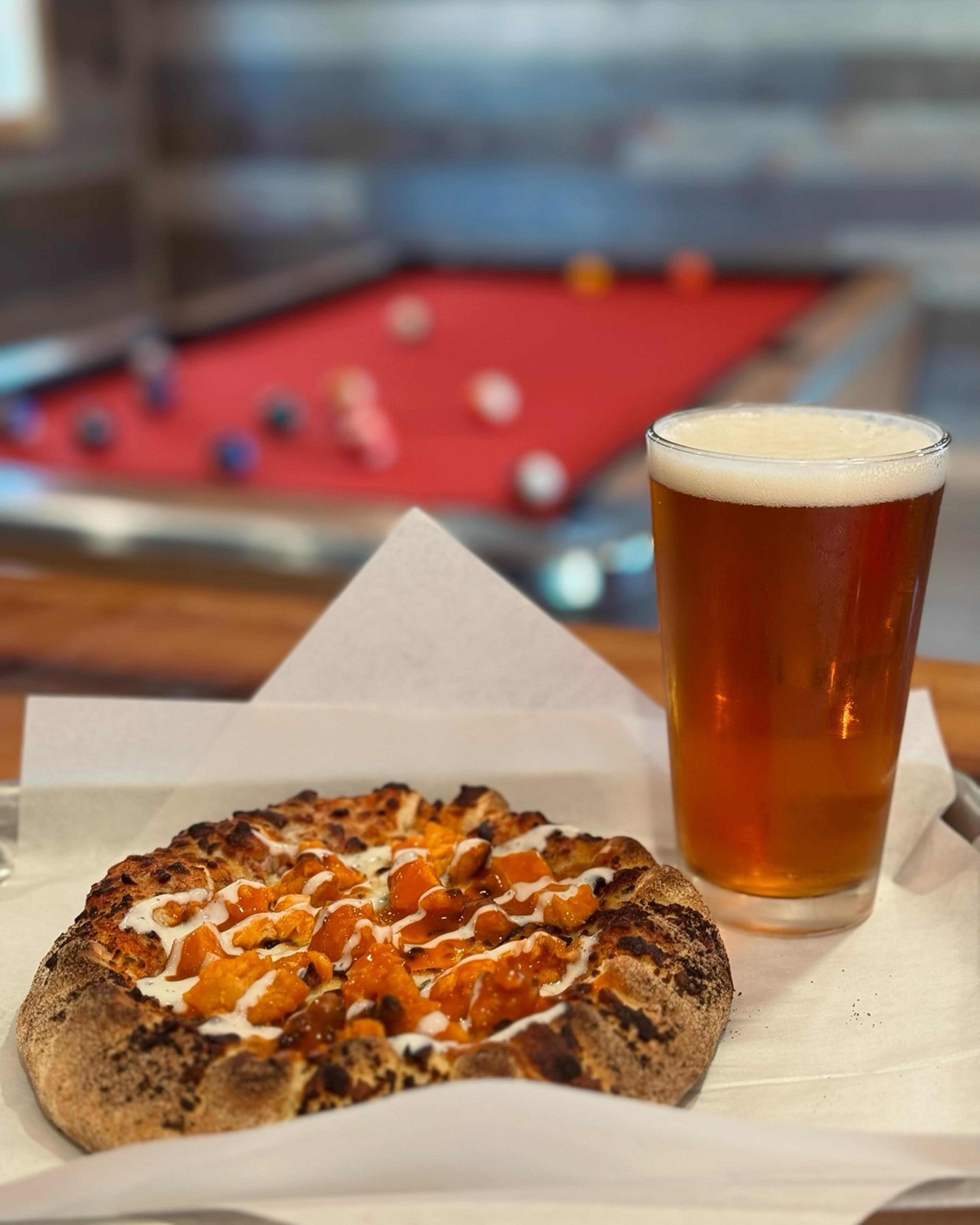 A pizza and glass of beer placed in front of a pool table.